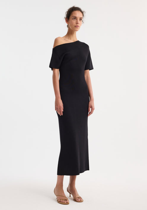 Asymmetrical knitted dress in black with one-shoulder design, fitted waist, and fluid midi skirt; model wears beige strappy sandals, hair pulled back.