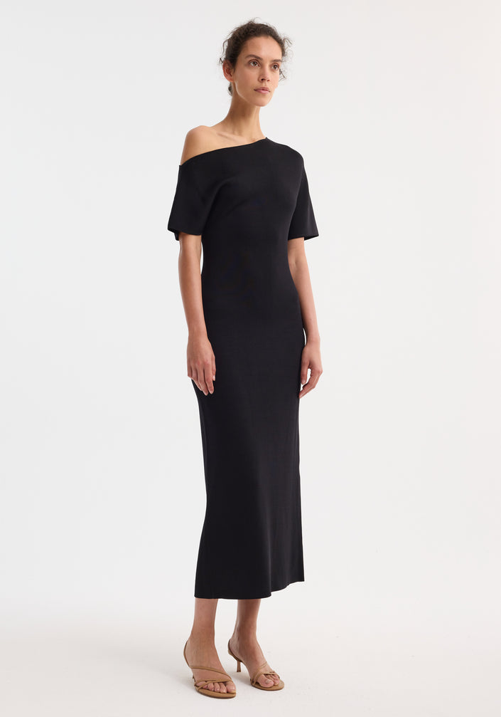 Asymmetrical knitted dress in black with one-shoulder design, fitted waist, and fluid midi skirt; model wears beige strappy sandals, hair pulled back.