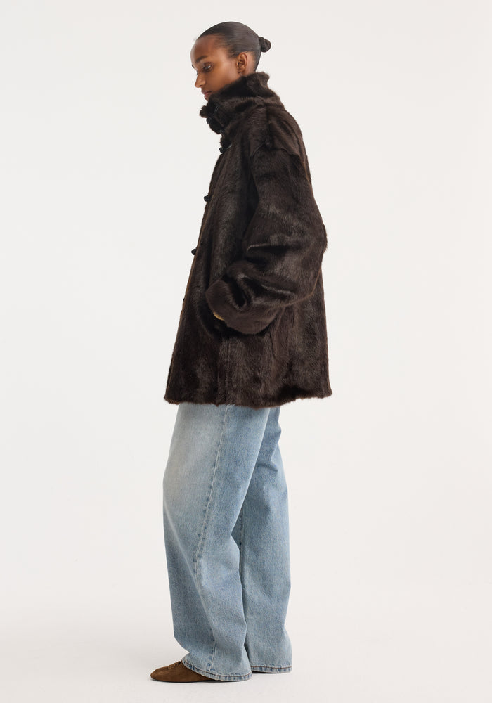 Person wearing Pankou closure faux fur short coat, dark brown, with toggle closures, stand-up collar, over light-washed jeans and pointed-toe flats, hair in bun.