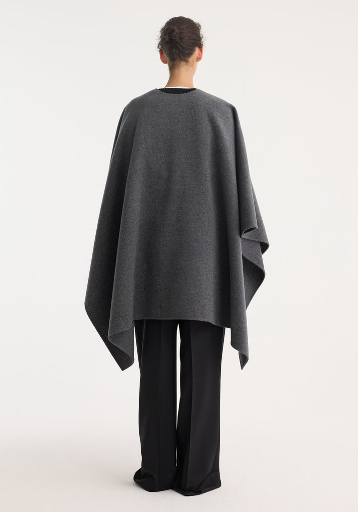 Back view of a person wearing a Double-faced belted poncho in mid grey melange, featuring an asymmetric drape over black trousers and dark shoes.