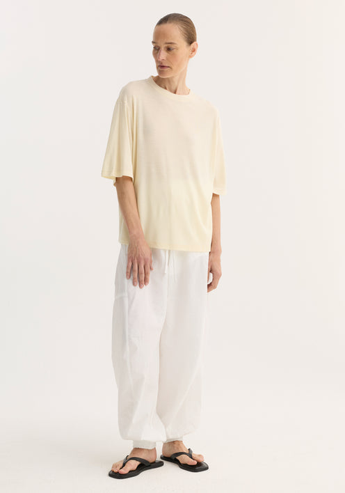 Model wearing an oversized merino t-shirt with a relaxed fit, dropped shoulders, and elbow-length sleeves, paired with white drawstring trousers and black sandals.