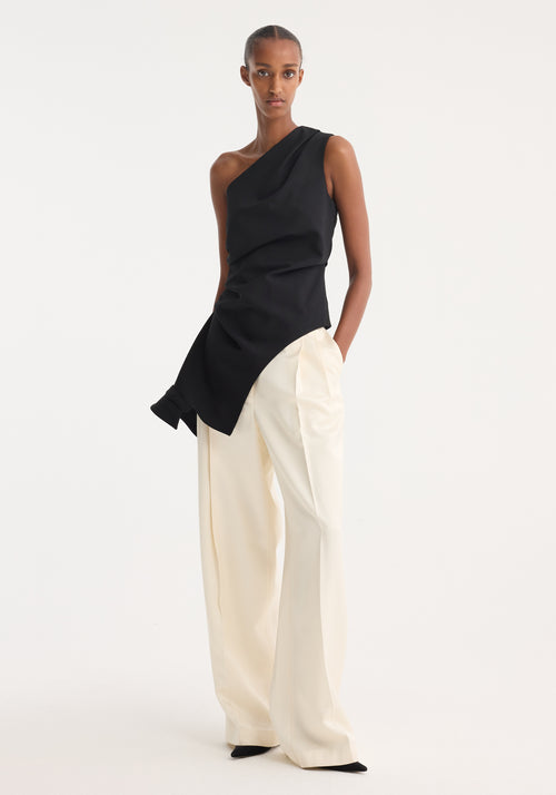 Model wearing Reimagined one shoulder top | black with asymmetrical design, draping detail, and fitted waist, paired with high-waisted cream trousers, hands in pockets.