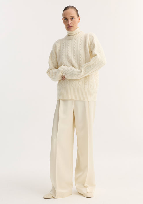 Person wearing an oversized, pure wool Cable knit jumper in off white with a round neckline and dropped shoulders, paired with wide-leg trousers and matching shoes.