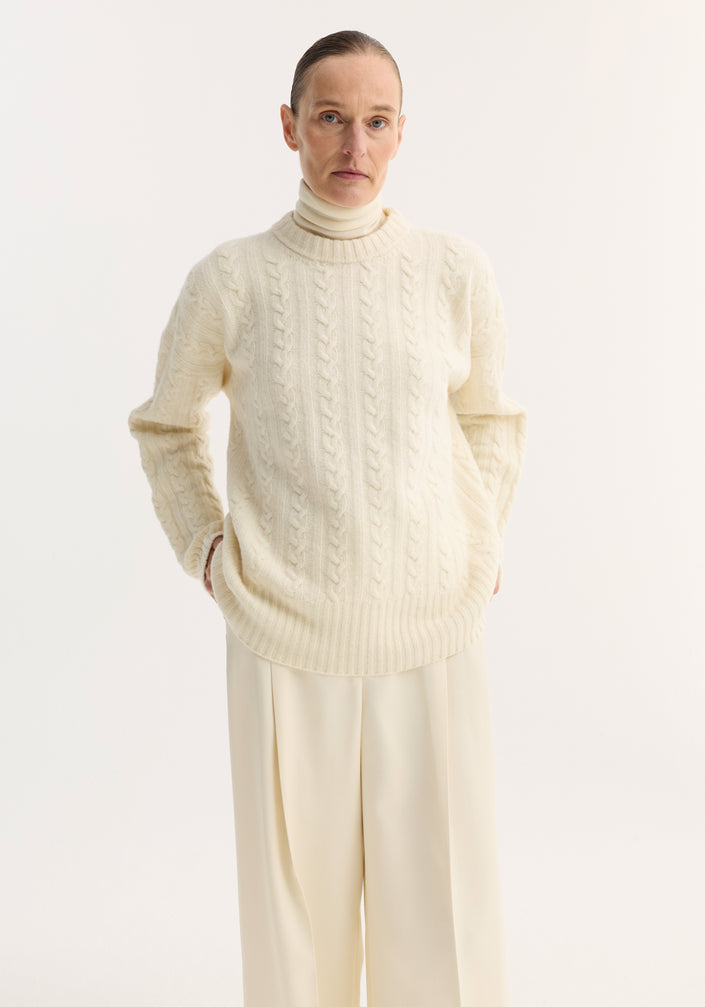 Person wearing an oversized, pure wool Cable knit jumper in off white with a round neckline and dropped shoulders, paired with wide-leg trousers and matching shoes.