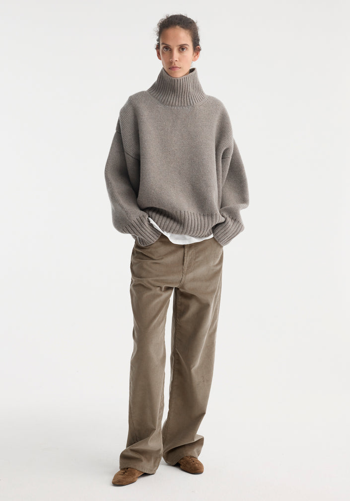 Person wearing Sculptural compact turtleneck | clay melange, an oversized grey rib-knit sweater over a white shirt, paired with light brown corduroy trousers and suede shoes.