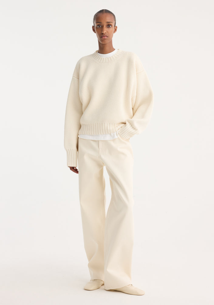 Person wearing a Sculptural compact crewneck in off white, a loose-fitting wool sweater with drop shoulders and a classic crewneck, paired with matching trousers and shoes.