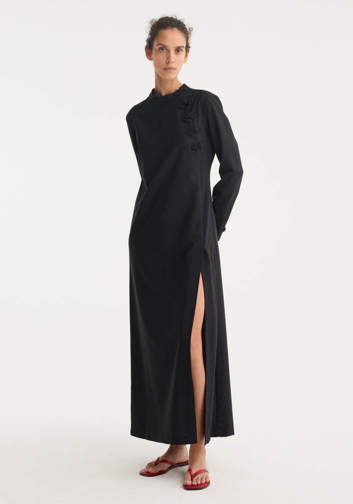 Model in Pankou closure dress: black, floor-length with long sleeves, side slit, and decorative closures on shoulder. Wears red sandals, hair pulled back.