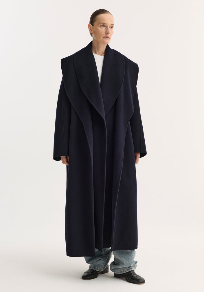 Model wears Signature double-faced scarf coat in navy, showcasing an oversized shawl collar, self-tie belt, and long silhouette over light-wash jeans and black shoes.