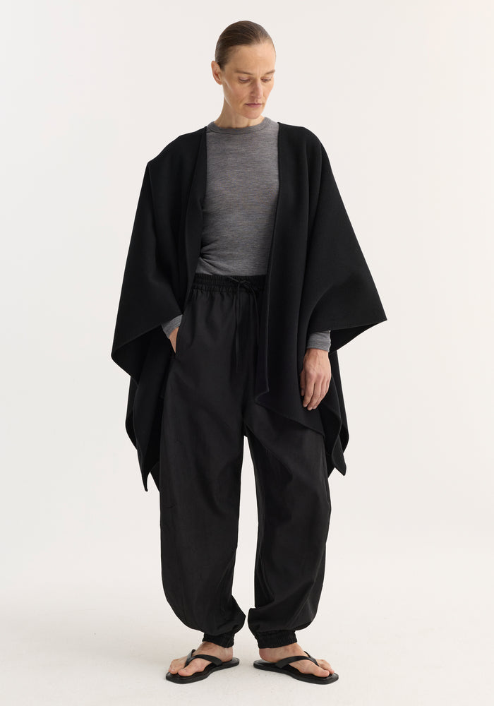 Person wearing Draped Scarf Poncho in black, showcasing its soft, flowing wool design over a grey top and black drawstring pants, with simple black sandals.