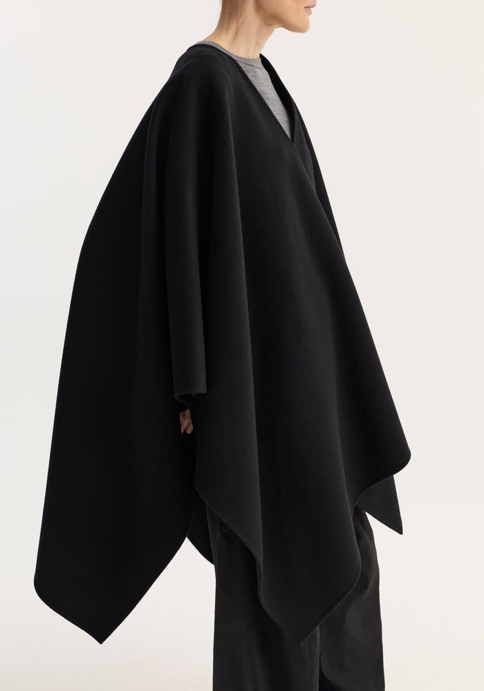 Side-profile of a person wearing the Draped scarf poncho | black, showcasing its voluminous, flowing silhouette made from soft, double-faced wool.