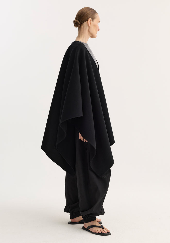 Model in a side-profile wears the Draped Scarf Poncho in black, showcasing its voluminous wool drape over a grey top, paired with loose black trousers.
