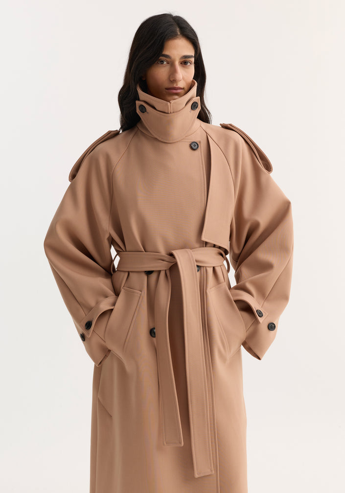 Woman wearing an oversized classic trench with structured detailing, wide collar, epaulettes, and cinched belt; black trousers peek from beneath, completing the modern look.