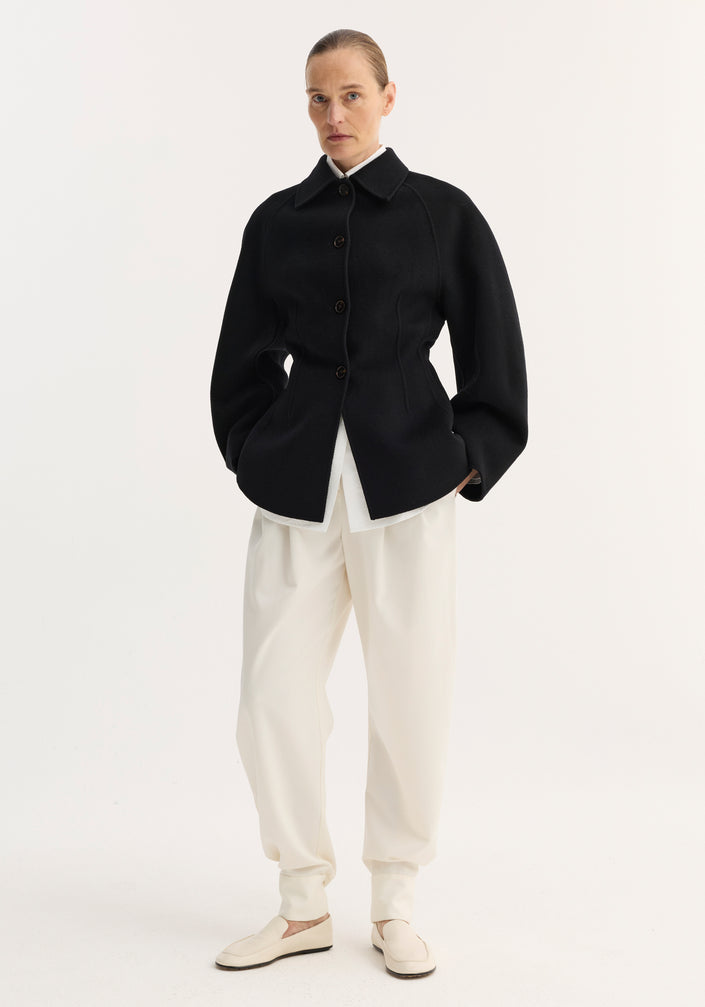 Woman wearing a double-faced sculptural jacket with button closures and sculpted silhouette, paired with ankle-gathered trousers and loafers, hair pulled back neatly.