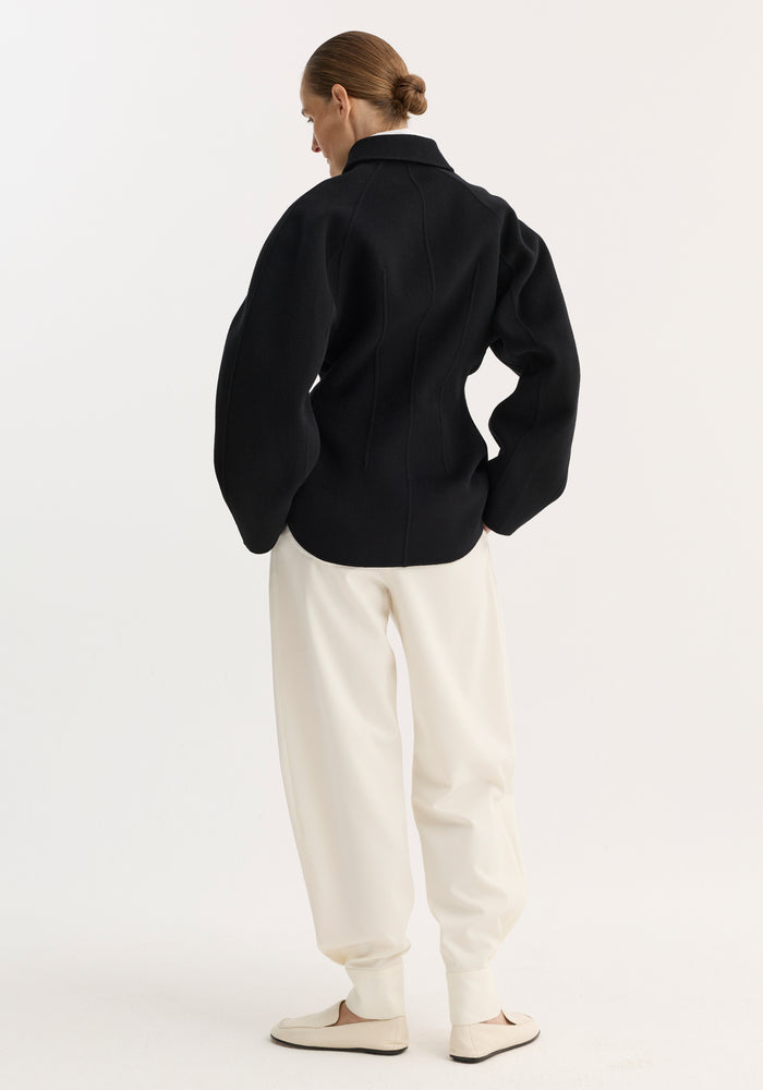 Model in double-faced sculptural jacket with voluminous sleeves, seaming, and button closures, paired with loose joggers and slip-ons, showcasing a tailored, architectural design.
