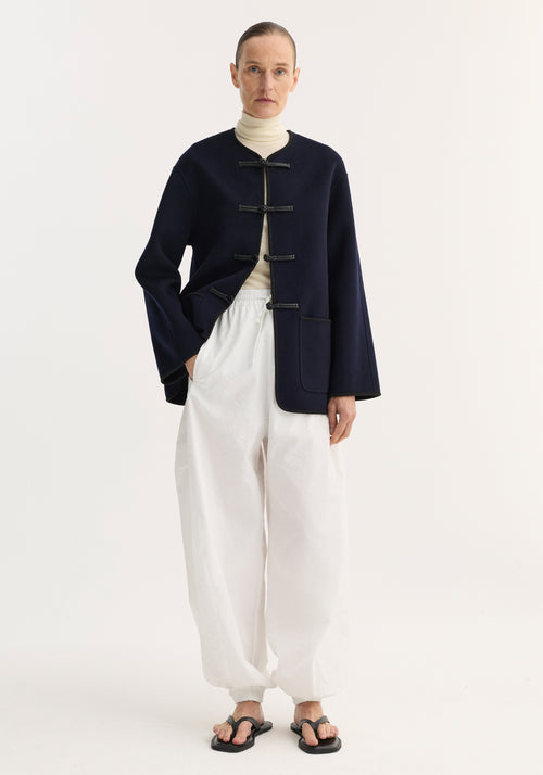 Person wearing the Mandarin double-faced wool jacket with toggle closures and patch pocket, layered over a cream turtleneck, paired with voluminous white trousers.