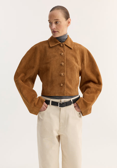Person in a Rounded textured suede jacket | cognac, featuring a cropped, waisted fit and structured sleeves, paired with a turtleneck, trousers, and loafers.