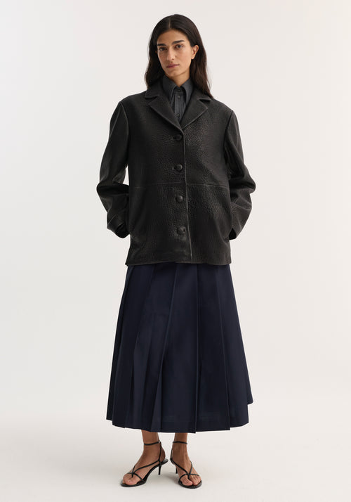 Model wearing a black Leather jacket with small lapel, featuring a textured finish, notched collar, button closures, and side pockets, styled over a dark shirt and navy pleated skirt.