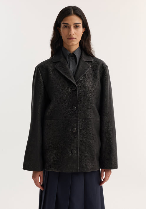 Model wearing a black Leather jacket with small lapel, featuring a textured finish, notched collar, button closures, and side pockets, styled over a dark shirt and navy pleated skirt.