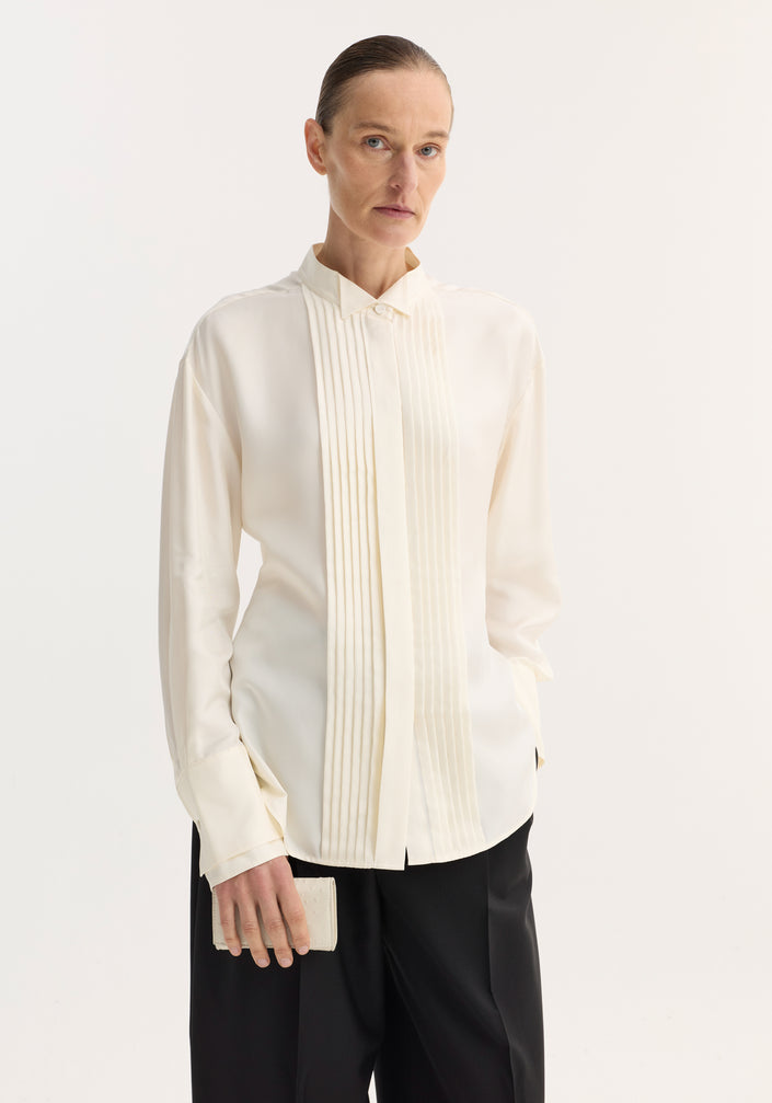 Model wearing Shaped Tuxedo Silk Shirt | Cream with stand-up collar and pleated front, paired with wide-leg trousers and holding an off-white clutch.