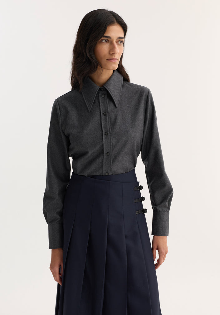 Woman in a fitted shirt, dark grey melange, with a sharp collar and button-down front, paired with a navy pleated midi skirt and strappy sandals.
