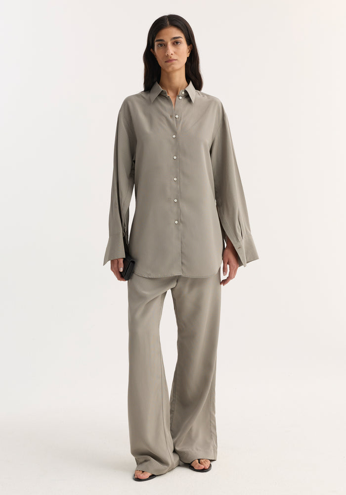 The woman models a Relaxed shaped shirt | light grey stripe, showcasing its loose fit, elongated sleeve plackets, and mother-of-pearl buttons, paired with matching trousers.