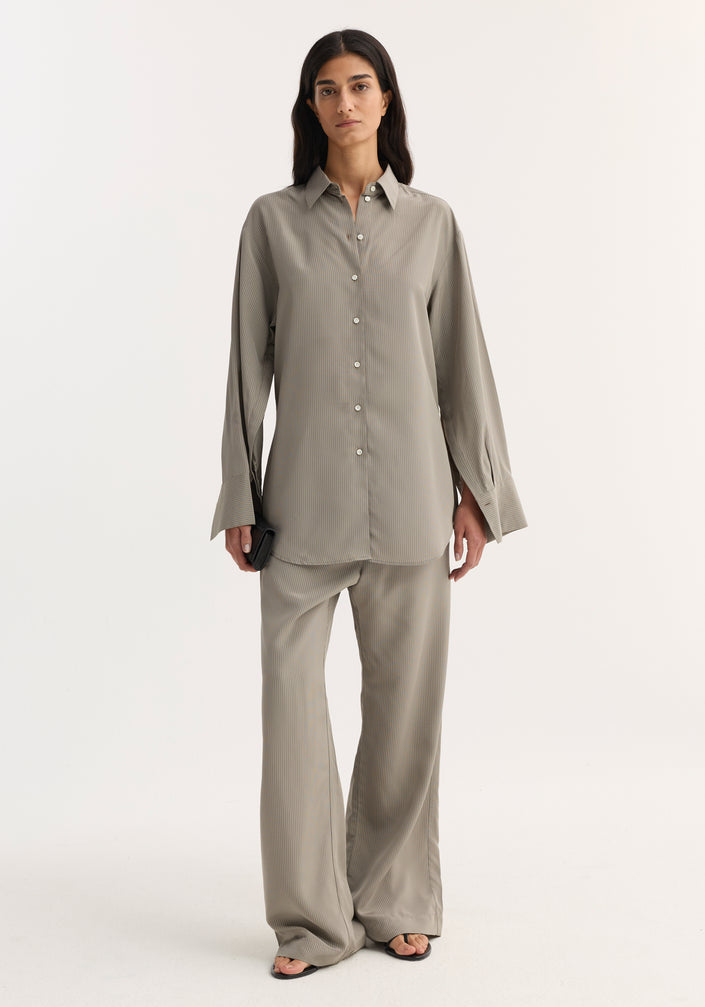 The woman models a Relaxed shaped shirt | light grey stripe, showcasing its loose fit, elongated sleeve plackets, and mother-of-pearl buttons, paired with matching trousers.