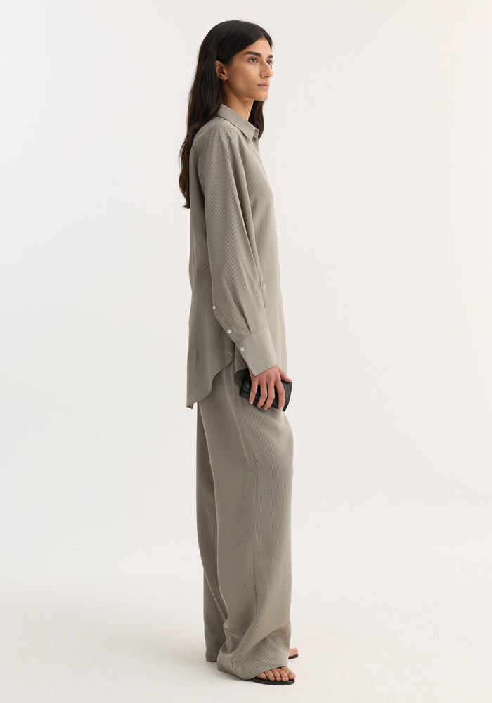 Person in profile wearing a Relaxed shaped shirt | light grey stripe, with extended sleeve plackets and rounded hem, paired with matching wide-leg trousers. Holding a black clutch.