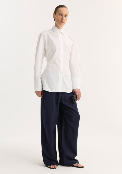 Person wearing Shaped shirt | optic white, a relaxed fit button-down with rounded hem, styled with navy trousers, black sandals, and a sweater over shoulders.
