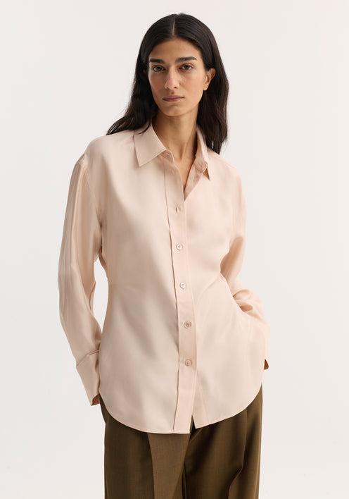 Silk shaped shirt in rose, featuring a relaxed hourglass silhouette with a button-down front and rounded hem, modeled with a knit sweater and wide-leg trousers.