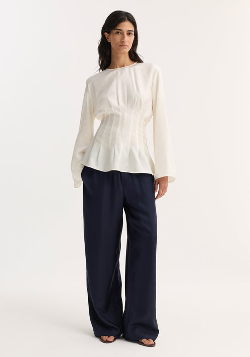 Woman wearing a Pleated Silk Top | Cream, featuring long sleeves, pleat details, and a back keyhole closure, paired with wide-leg navy trousers and open-toe sandals.