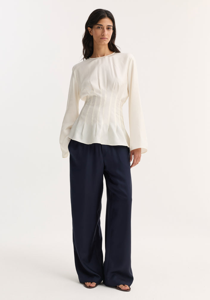 Woman wearing a Pleated Silk Top | Cream, featuring long sleeves, pleat details, and a back keyhole closure, paired with wide-leg navy trousers and open-toe sandals.