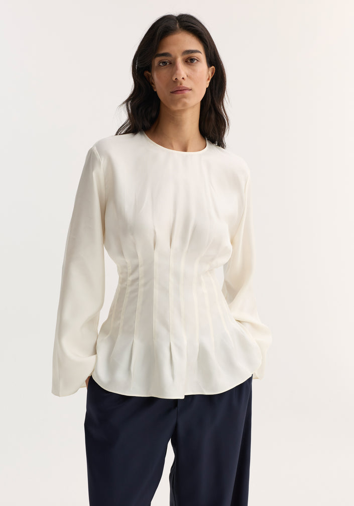 Woman wearing a Pleated Silk Top | Cream, featuring long sleeves, pleat details, and a back keyhole closure, paired with wide-leg navy trousers and open-toe sandals.