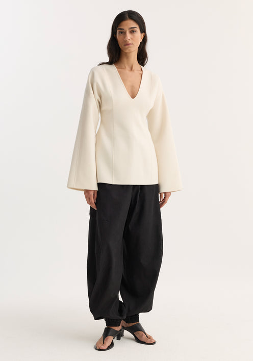 Woman wearing Sculptural long sleeve top in off white with voluminous sleeves and a defined waist, paired with relaxed black pants and heeled sandals.