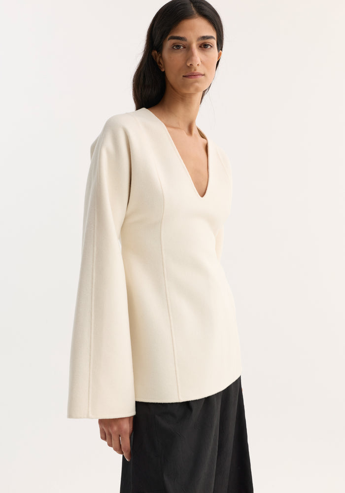 Woman modeling Sculptural long sleeve top | off white, showcasing its voluminous sleeves, defined waist, and V-neckline, paired with black wide-leg trousers.