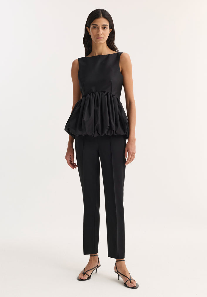 Model wearing a Voluminous boat-neck top in black, showcasing a sleeveless design with a sculptural peplum hem, paired with tailored trousers and strappy sandals.