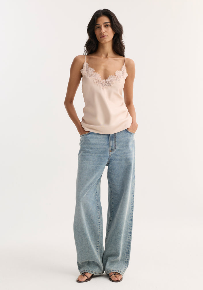 Woman wearing a lace silk camisole in rose with lace-trimmed V-neck and adjustable straps, paired with wide-leg jeans, standing with hands in pockets.