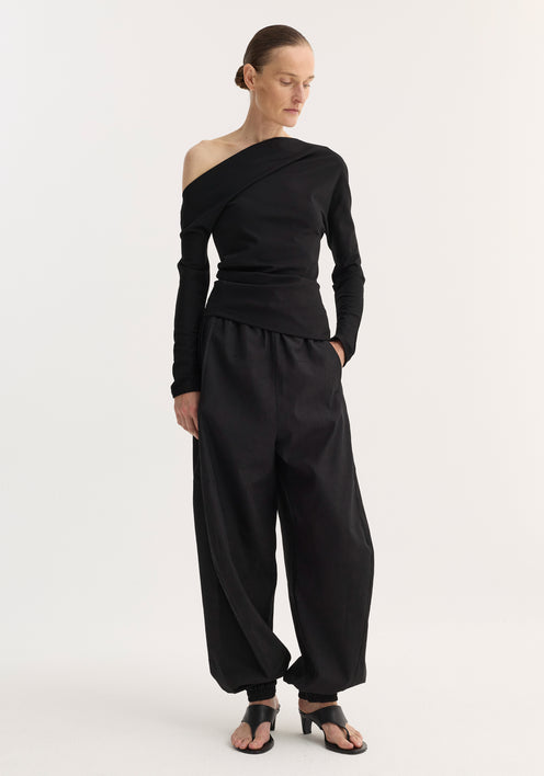 Woman in black Twisted jersey top with asymmetrical neckline and long sleeves, paired with harem pants and heeled sandals, exuding a modern, minimal style.