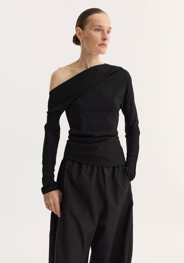 Woman in black Twisted jersey top with asymmetrical neckline and long sleeves, paired with harem pants and heeled sandals, exuding a modern, minimal style.