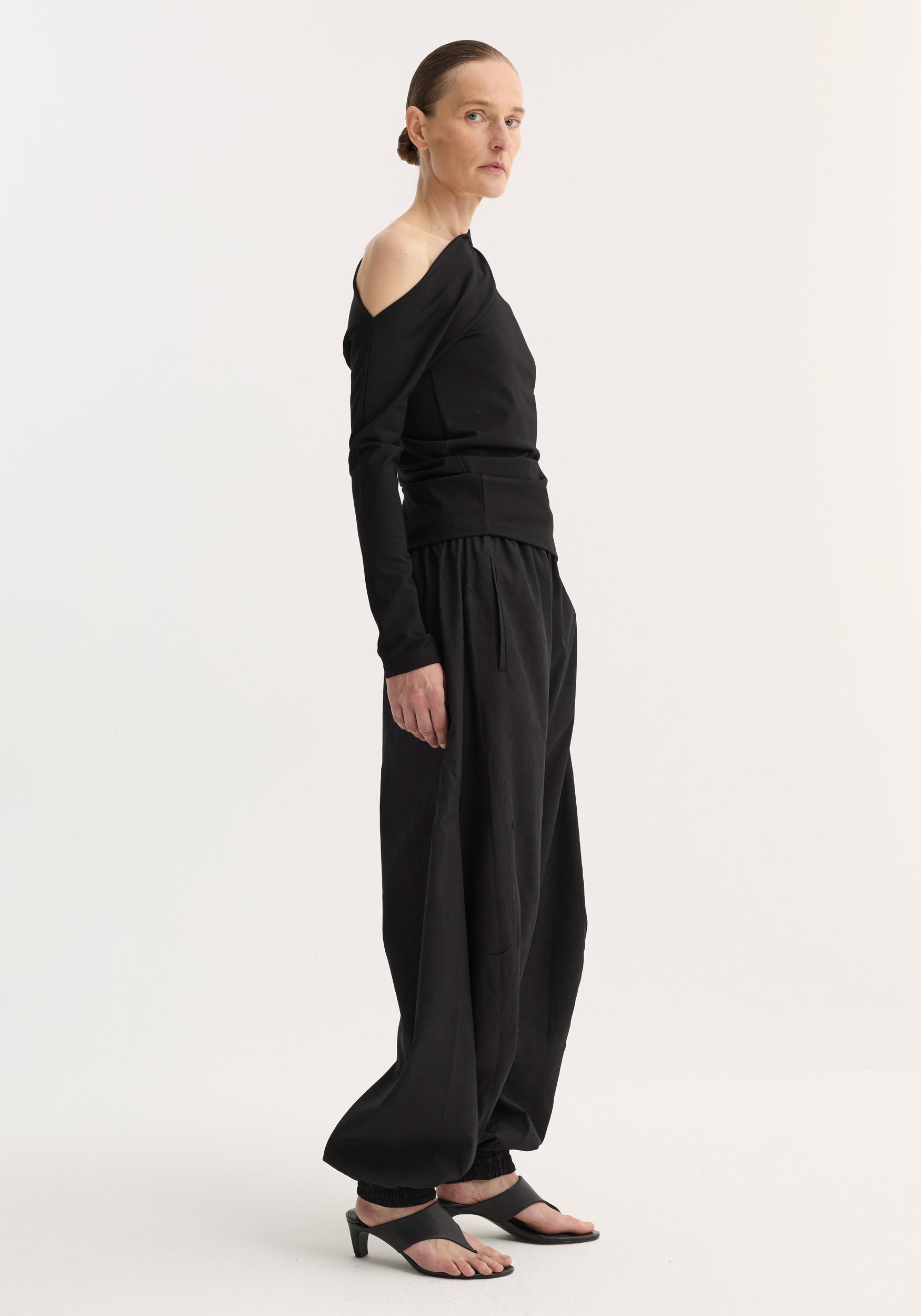 Woman in profile wearing a Twisted jersey top; black, one-shoulder, long-sleeve with draped waist detail, paired with loose trousers and open-toe sandals.