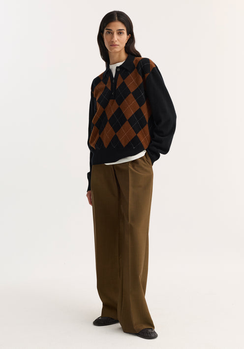 Person wearing a Compact knitted check polo in black rust check with argyle pattern, layered over a white top, paired with high-waisted trousers and slip-on shoes.