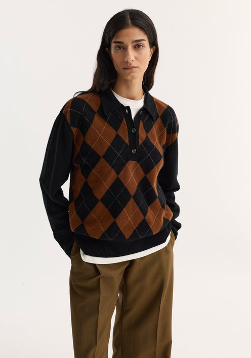 Person wearing a Compact knitted check polo in black rust check with argyle pattern, layered over a white top, paired with high-waisted trousers and slip-on shoes.