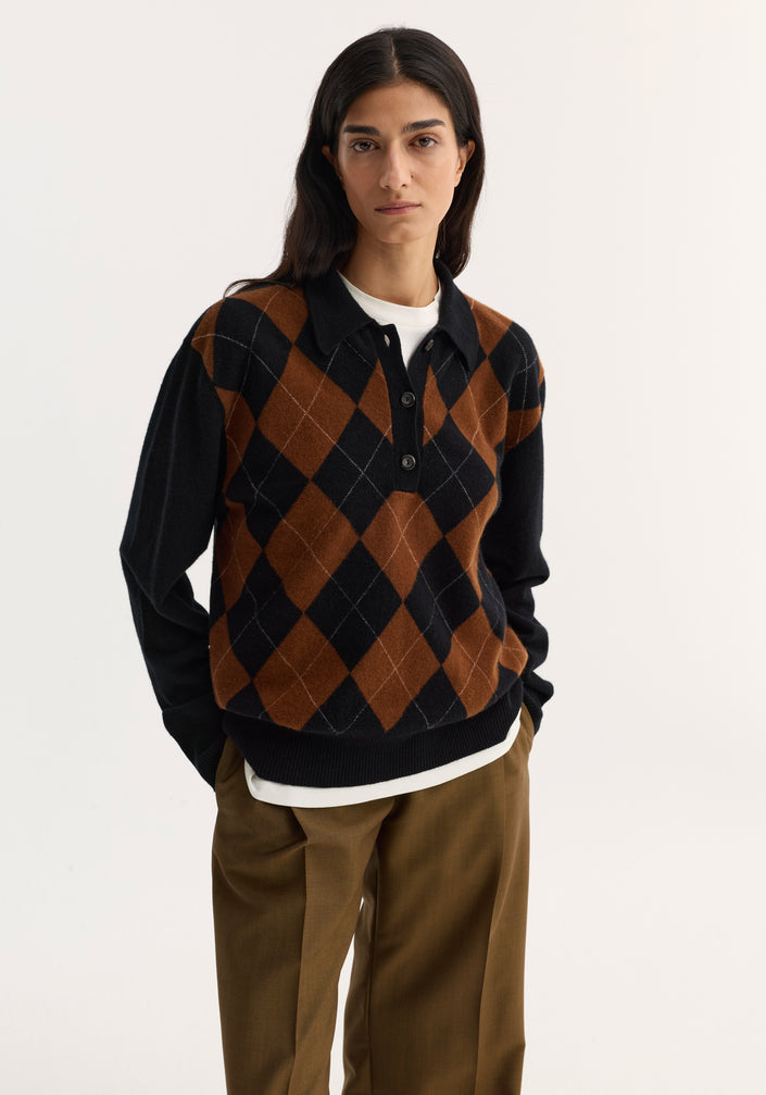 Person wearing a Compact knitted check polo in black rust check with argyle pattern, layered over a white top, paired with high-waisted trousers and slip-on shoes.