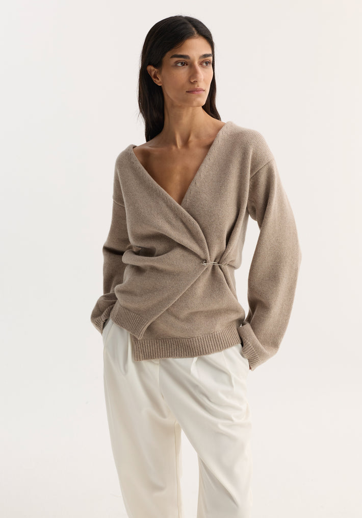 A model wears an off-shoulder wrap cardigan in light taupe, featuring a relaxed fit and metal safety-pin closure, paired with high-waisted, wide-leg trousers.