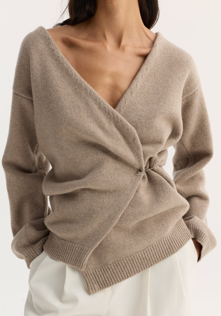 Person wearing off-shoulder wrap cardigan in light taupe, wool-cashmere blend, with safety pin closure and ribbed hem, paired with off-white trousers.