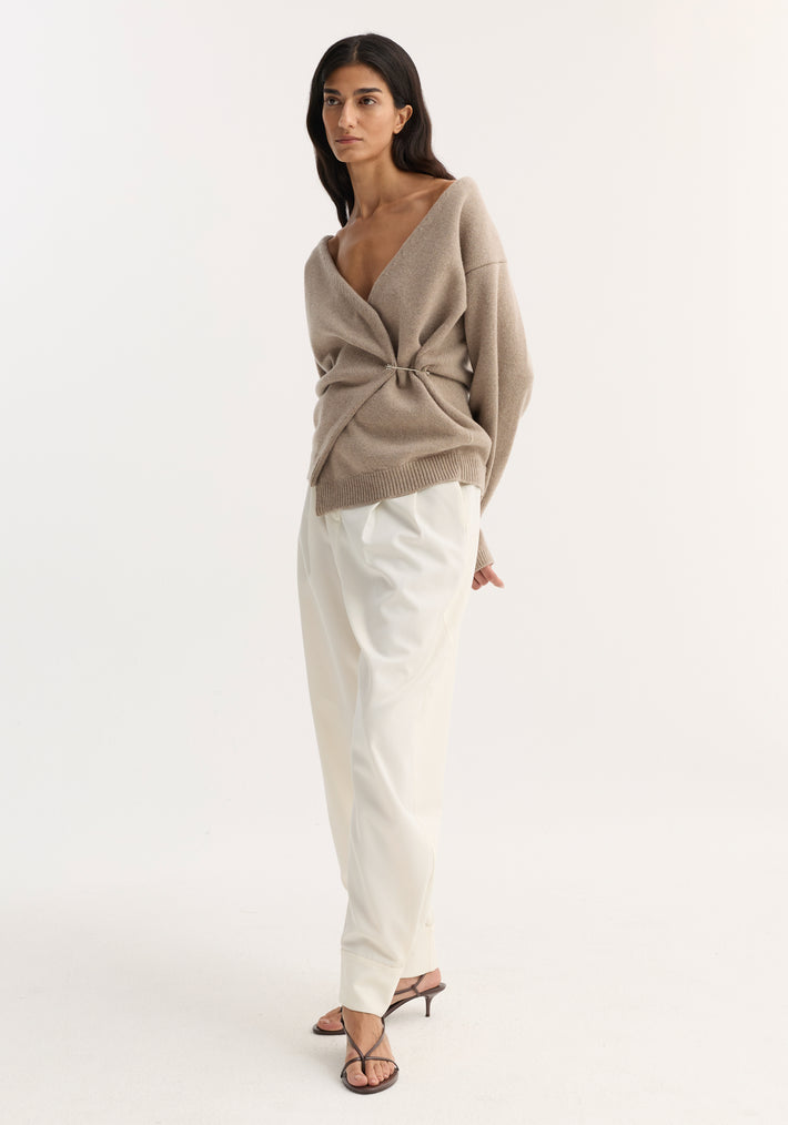 Woman in a light taupe off-shoulder wrap cardigan with metal clasp, paired with high-waisted white trousers and dark brown strappy heeled sandals.