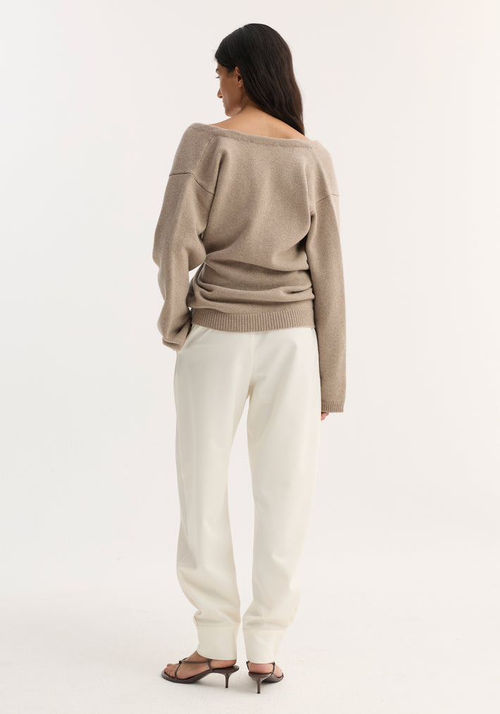 Woman in an off-shoulder wrap cardigan, light taupe, soft wool-cashmere blend, secured with metal safety pin, paired with loose ivory trousers and dark sandals.