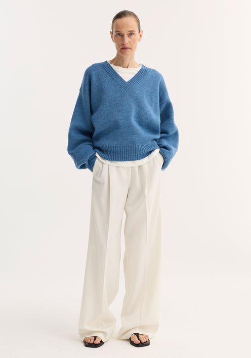 Model wearing a denim blue melange V-neck knitted jumper with a relaxed fit, layered over a white top, paired with wide-leg off-white trousers.