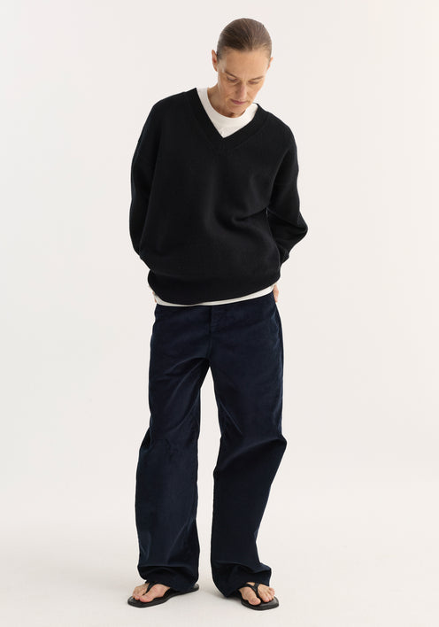 Model wearing V-neck knitted jumper in black, featuring a relaxed fit, drop shoulders, over a white T-shirt with wide-leg corduroy trousers and flip-flop sandals.