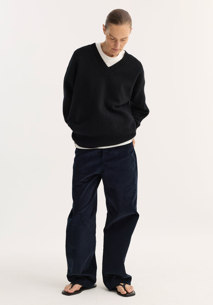 Model wearing V-neck knitted jumper in black, featuring a relaxed fit, drop shoulders, over a white T-shirt with wide-leg corduroy trousers and flip-flop sandals.