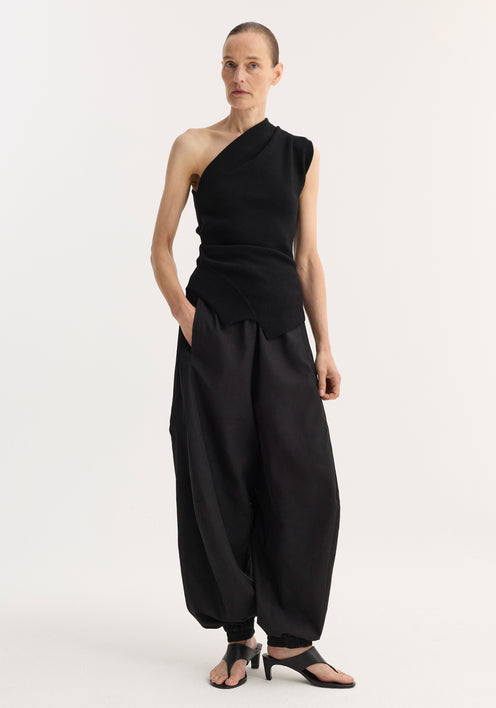 Off-shoulder top | black: Model wears a black, one-shoulder knit top with an asymmetrical hem and fitted waist, paired with harem-style trousers and open-toe sandals.